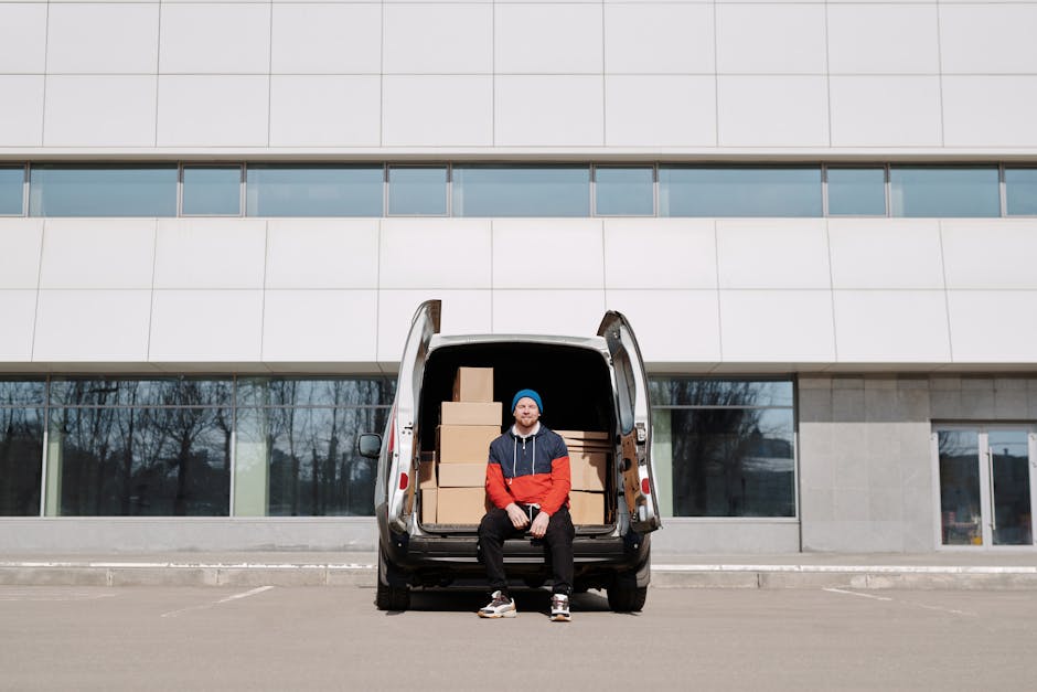 A man wearing a multicoloured jacket with blue, red, and black sections, along with a blue beanie, is seated on the open tailgate of a black van parked on a paved area outside a modern, multi-story bu