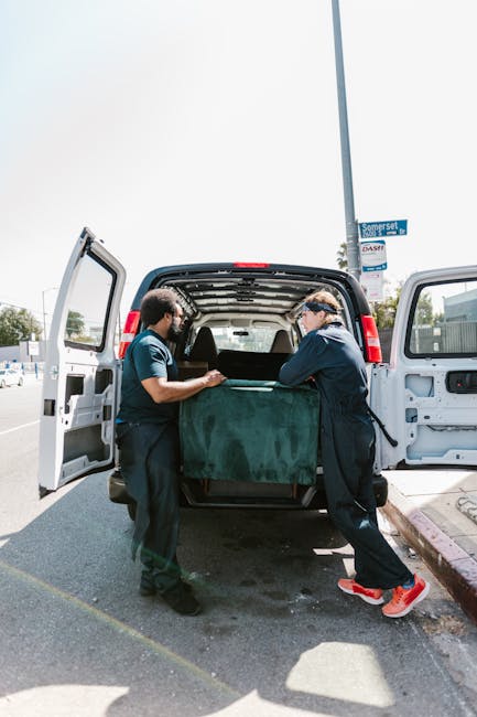 A man wearing a multicoloured jacket with blue, red, and black sections, along with a blue beanie, is seated on the open tailgate of a black van parked on a paved area outside a modern, multi-story bu