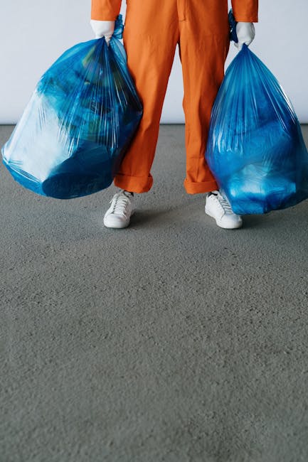 A person wearing a yellow rubber glove is reaching into an open cardboard box filled with various batteries, including AA, AAA, and 9V types, some with visible branding such as Power and Energizer. Th