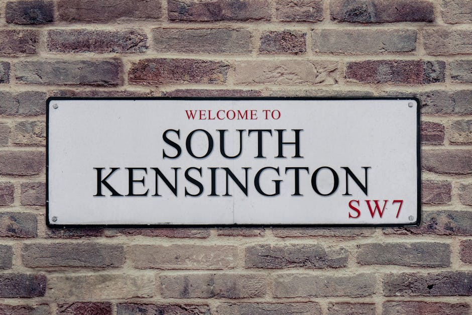 A narrow cobblestone street in Earl Court, South Kensington, lined with white residential buildings on both sides. The buildings feature large windows, some with black wrought iron railings, and small