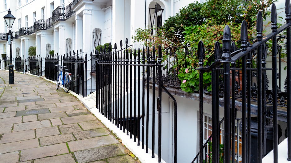 A paved sidewalk runs alongside a row of white terraced houses with black wrought iron railings and gates. A blue bicycle is parked and secured to one of the railings near the entrance of a basement flat. The houses feature large windows with black metal decorative covers and small balconies. Green foliage and flowering plants are visible on the terraces, with sunlight illuminating the scene. The image depicts a residential area suitable for house removals and furniture transport, with the outdoor environment prepared for moving logistics by a professional removals company such as South Kensington removals experts for SW7 flats.