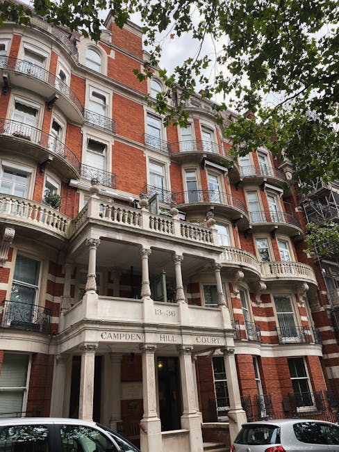 Exterior view of a multi-storey red brick residential building with ornate white stone balconies and decorative columns at the entrance, located at 13-36 Campden Hill Court. The building features multiple windows, some with flower boxes, and is partially obscured by overhanging green tree branches. In the foreground, several parked cars are visible on the street pavement. This scene illustrates the type of property involved in house removals and relocation services offered by the company, highlighting the building's classic architecture typical of South Kensington, with the entrance area prepared for a furniture transport or home relocation process.